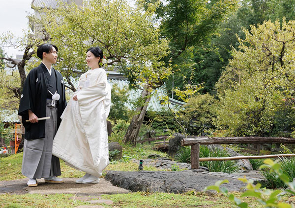 日本庭園で仲睦まじく見つめ合う、白無垢の花嫁と紋付袴の新郎の全身写真。