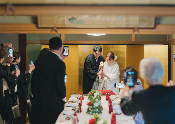 披露宴会場で紋付袴の新郎と白無垢の新婦が鏡開きを行う様子。周囲ではゲストがスマートフォンでその瞬間を撮影している