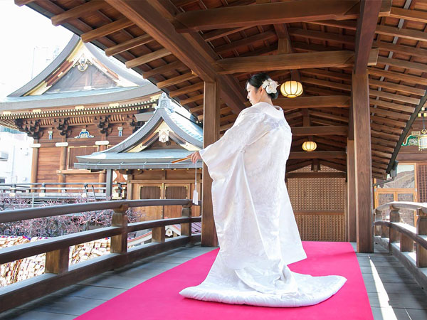 朱色の敷物が敷かれた社殿の回廊に立つ、白無垢姿の花嫁の後ろ姿