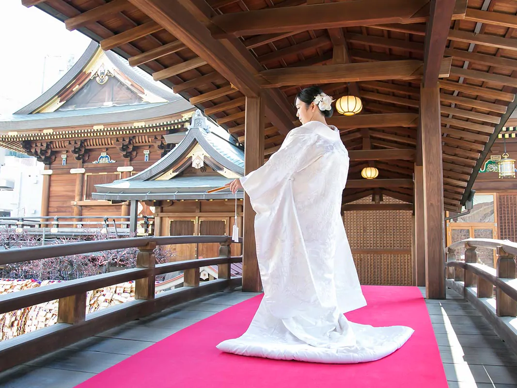 朱色の敷物が敷かれた社殿の回廊に立つ、白無垢姿の花嫁の後ろ姿