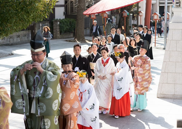 赤い和傘の下、神職と巫女に先導されて参道を進む花嫁と参列者の列