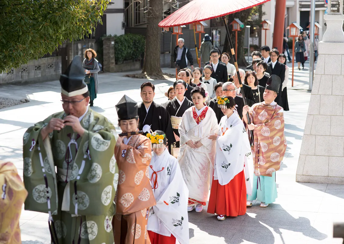 赤い和傘の下、神職と巫女に先導されて参道を進む花嫁と参列者の列