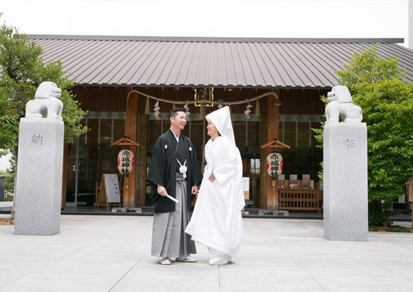 狛犬が並ぶ社殿前で向かい合う、綿帽子に白無垢の新婦と紋付袴の新郎