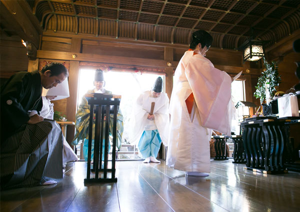 社殿の中で神職が祝詞を奏上し、頭を下げる紋付袴の新郎と白無垢の新婦