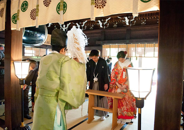 白い幕が下がる社殿で、神職の後ろ姿の先に頭を下げる紋付袴の新郎と色打掛の新婦