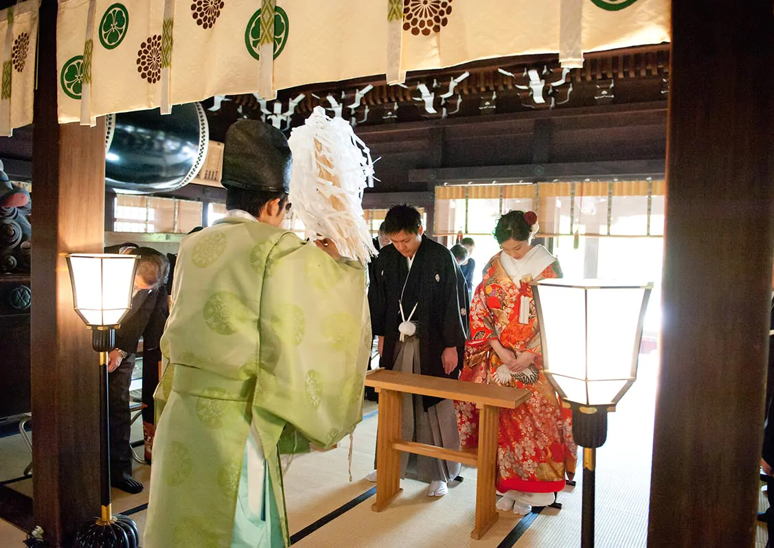 白い幕が下がる社殿で、神職の後ろ姿の先に頭を下げる紋付袴の新郎と色打掛の新婦