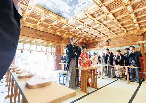 格天井の社殿で三々九度の盃を口に運ぶ紋付袴の新郎と、隣に立つ色打掛の新婦