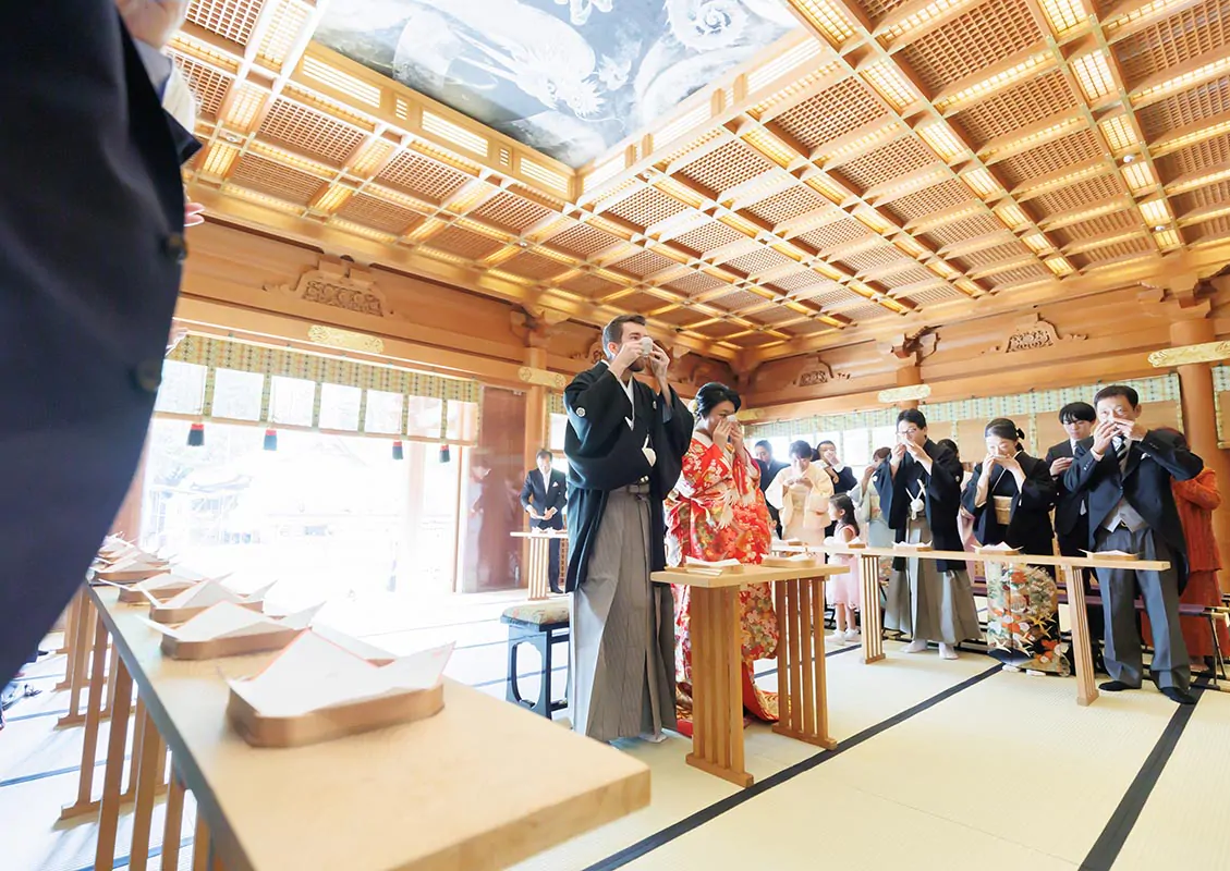 格天井の社殿で三々九度の盃を口に運ぶ紋付袴の新郎と、隣に立つ色打掛の新婦