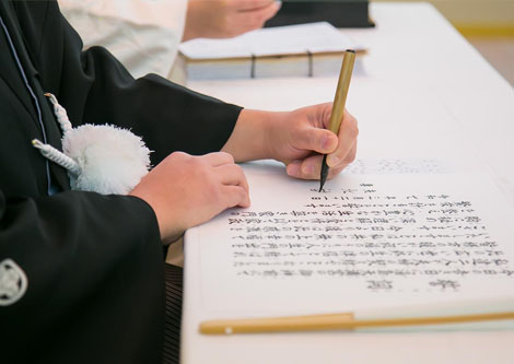 黒い紋付姿の神職が巻紙に毛筆で祝詞を書き記す手元
