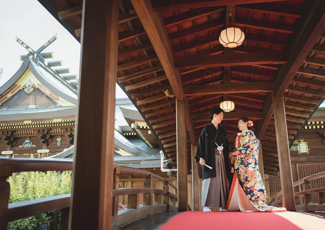 提灯が下がる神社の木造の回廊で、赤い毛氈の上に立つ色打掛の花嫁と羽織袴の新郎