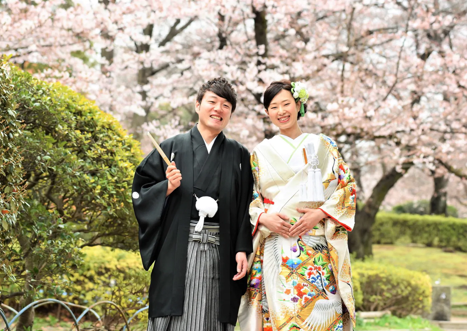 満開の桜並木を背に、鶴柄の白地の色打掛の花嫁と羽織袴の新郎が笑顔で並ぶ