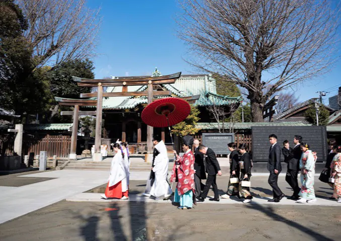 青空の下、鳥居の前を朱色の和傘を差しかけられた花嫁と新郎、巫女、家族が列になって進む参進