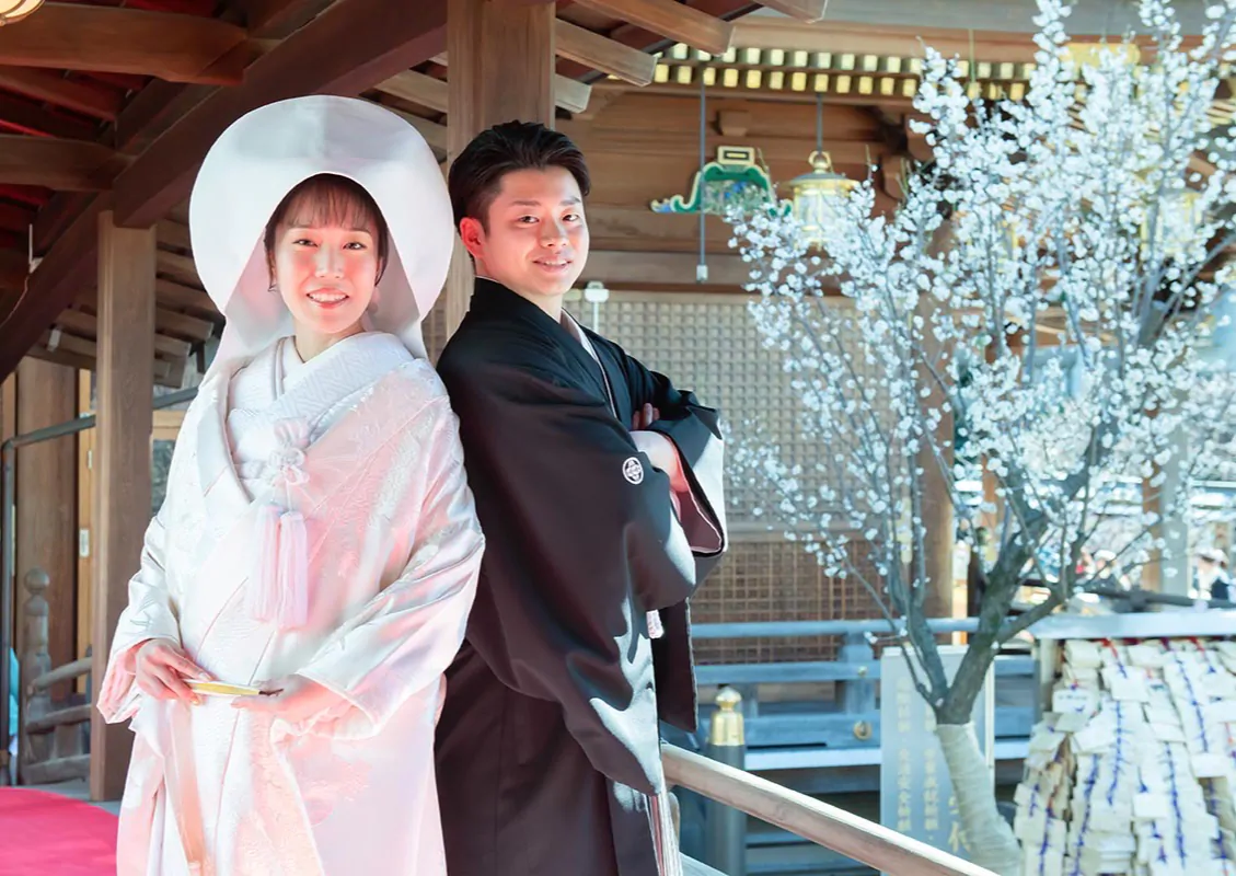白い梅の花が咲く社殿の廊下で、白無垢に綿帽子の花嫁と黒紋付の新郎が背中合わせで微笑む