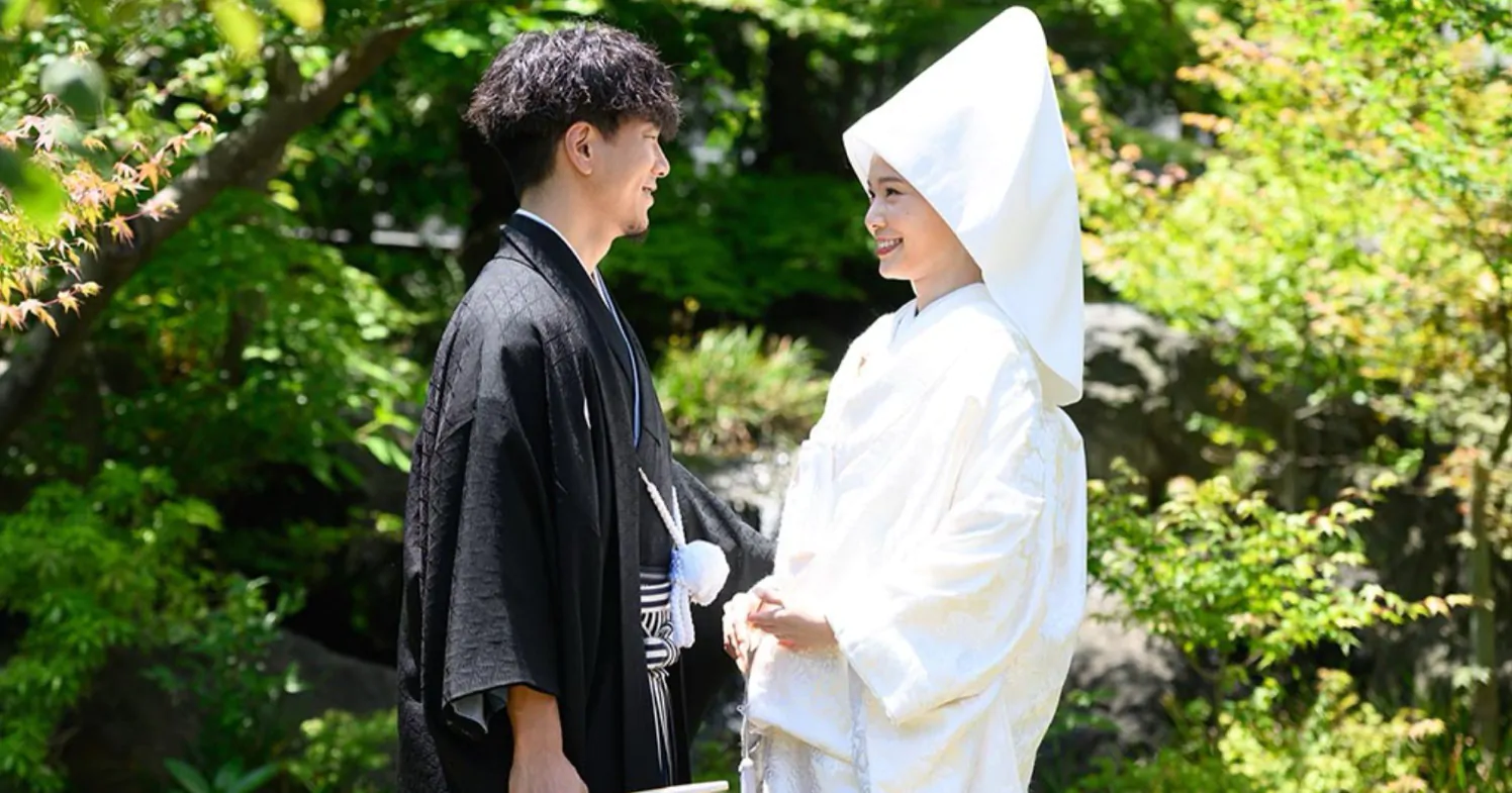 新緑の庭園で、白無垢に綿帽子の花嫁と黒紋付羽織袴の新郎が向かい合って微笑む